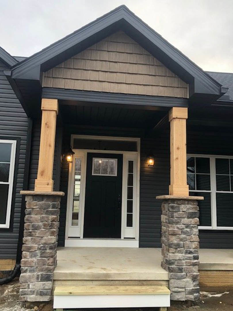 Front porch with stone pillars topped in black, black front door with glass window, stone ledge beneath window, gray siding, outdoor stairs leading to entrance
