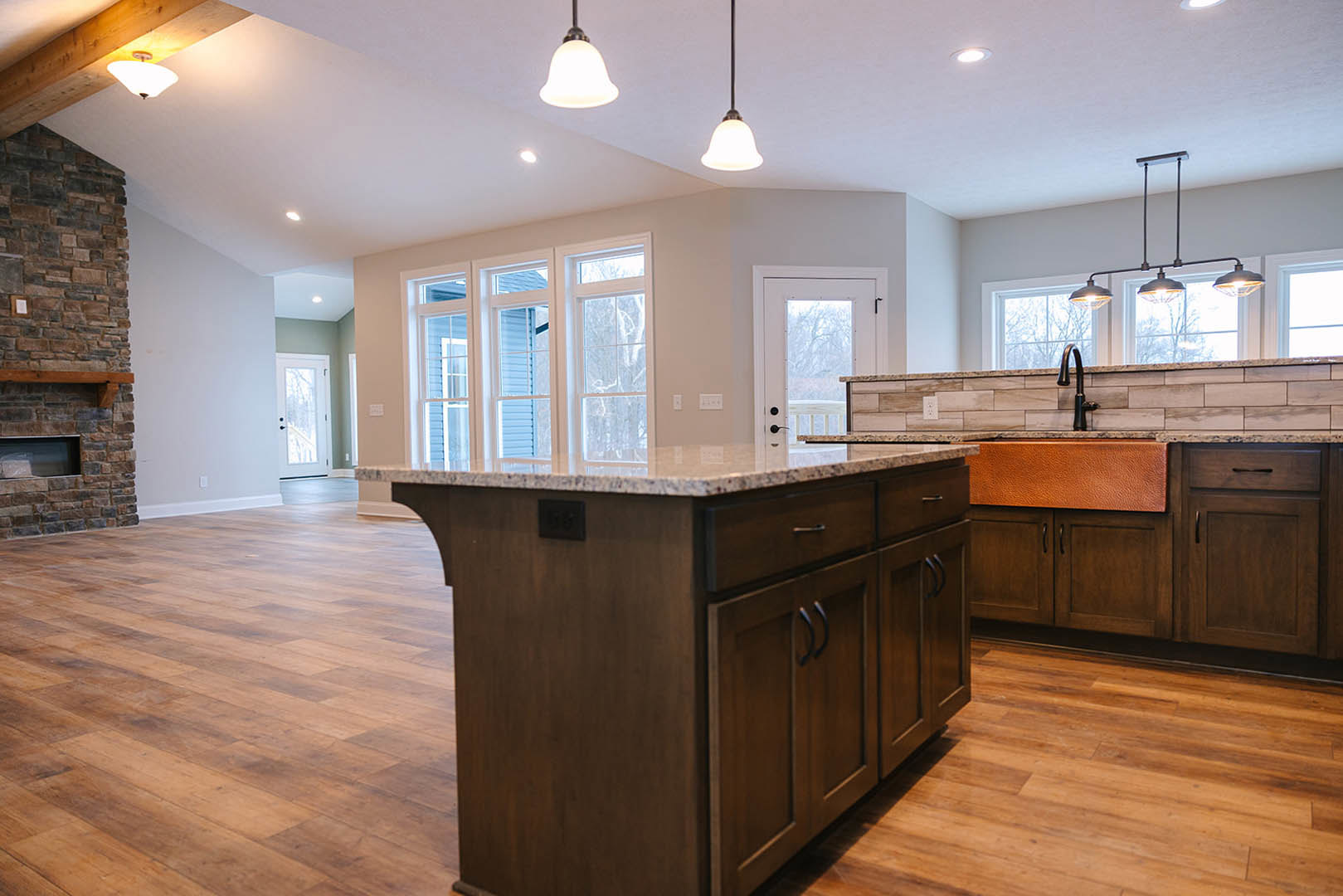 Granite kitchen island with white cabinetry, pendant light fixtures overhead, stone fireplace with wooden mantel, hardwood flooring, and large window in background