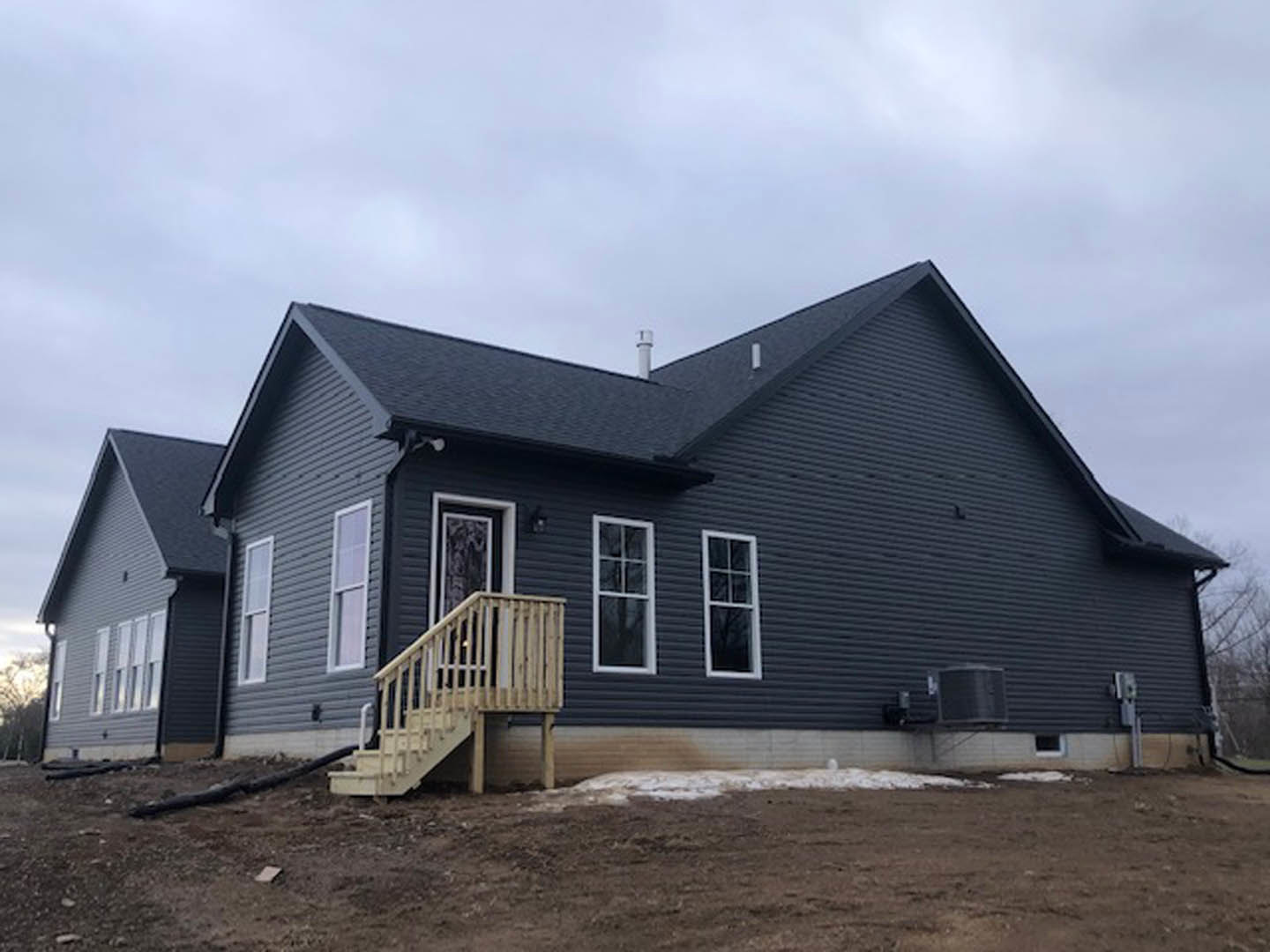 Two-story home with light-colored siding, large white-framed windows, and an exterior wooden staircase leading to the upper level, set against a cloudy sky with dirt landscaping in