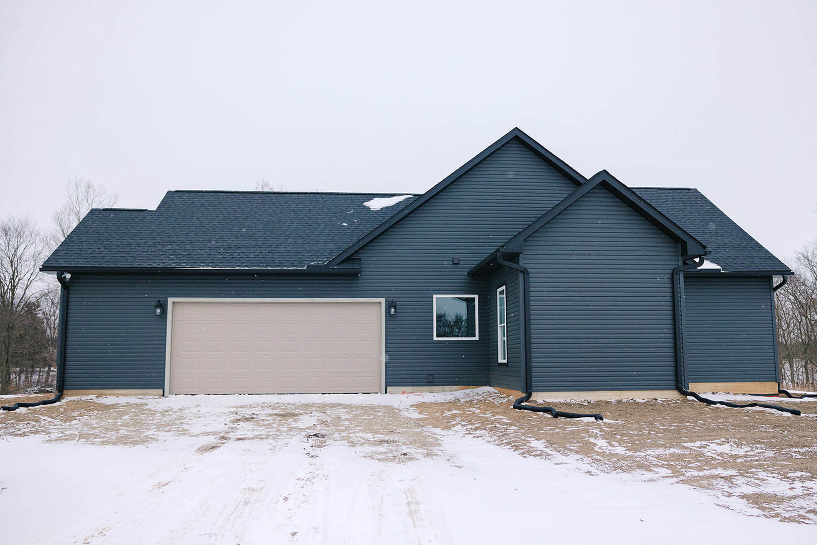 White garage door and blue roof on a house, snow covering the driveway with visible tire tracks, siding and window framed by bare trees in the background.