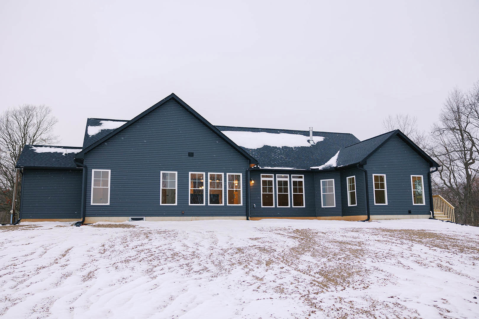 Two-story cottage with white-trimmed windows, snow-covered roof and ground, bare trees, and wooden staircase visible near entry