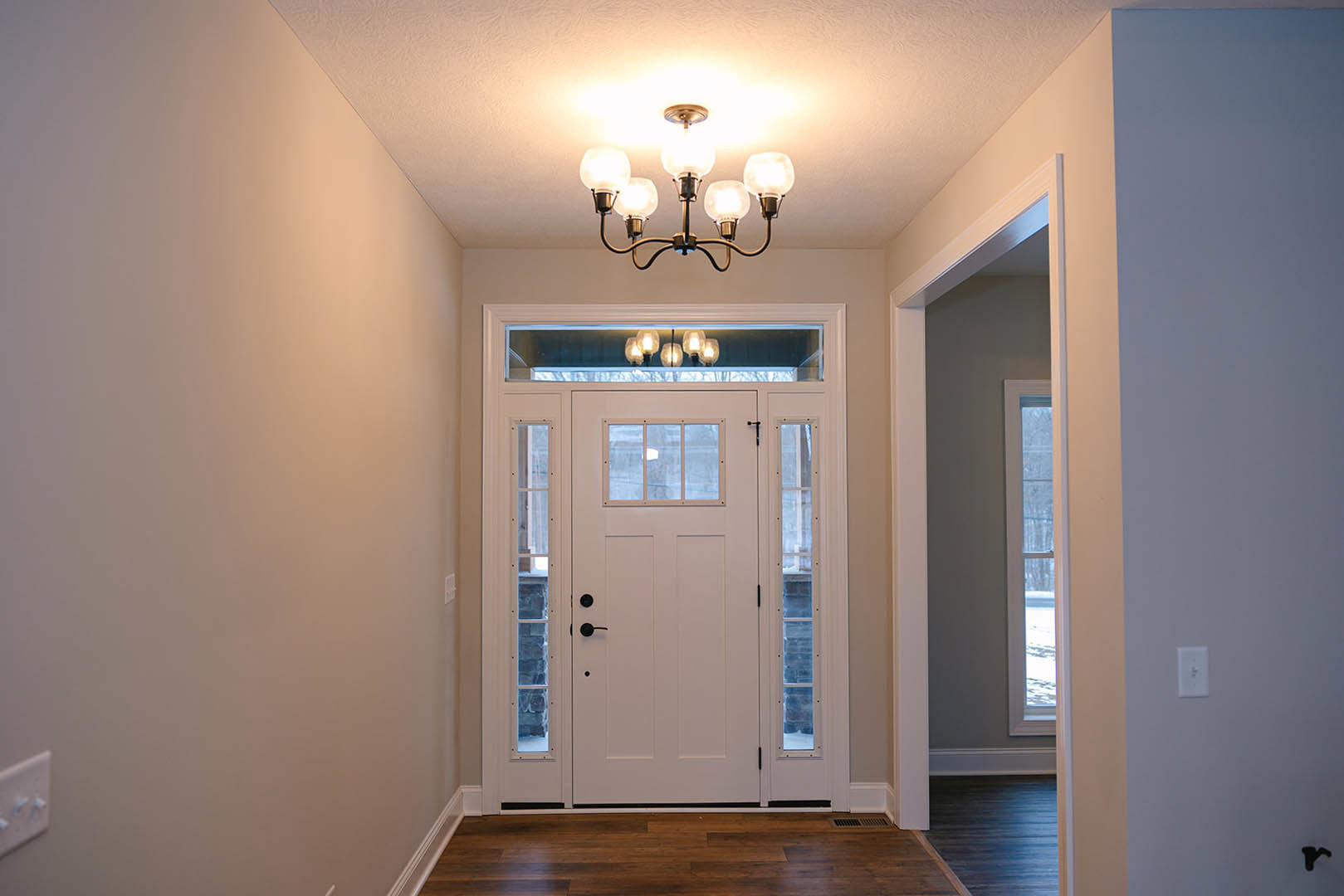 Chandelier with glass accents hanging from white hallway ceiling, dark wood flooring with white baseboard, white door with glass panes, light switch on wall