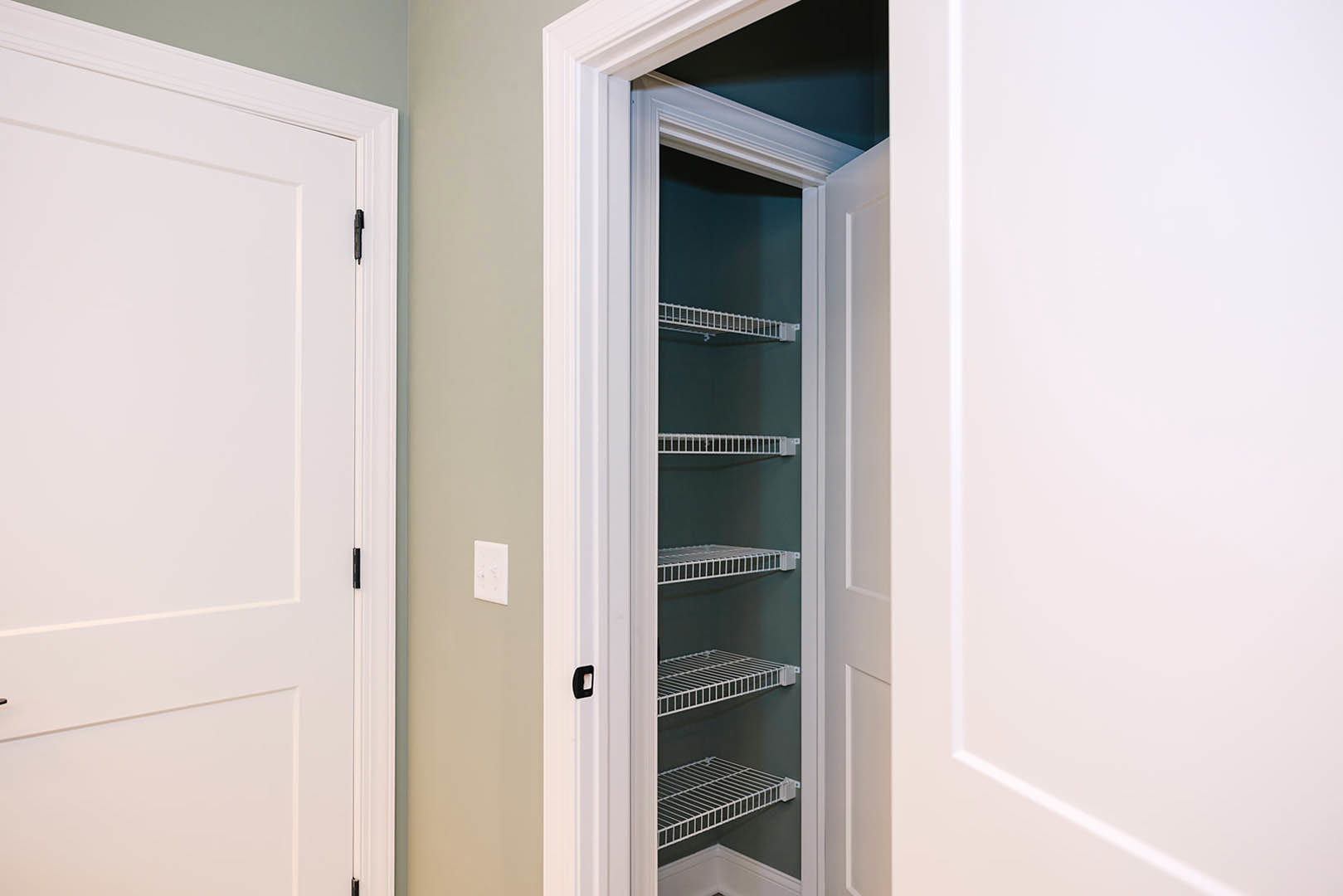Closet interior with white built-in shelves, white door featuring black hinges, and metal shelving unit with white bars against a dark wall