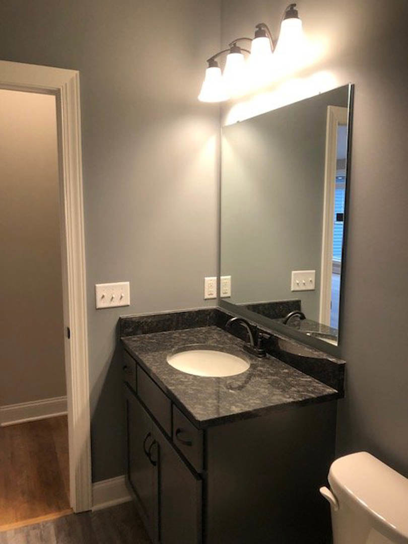 Modern bathroom with rectangular mirror above white sink, black countertop, chrome faucet, white tile backsplash, and row of wall-mounted light fixtures
