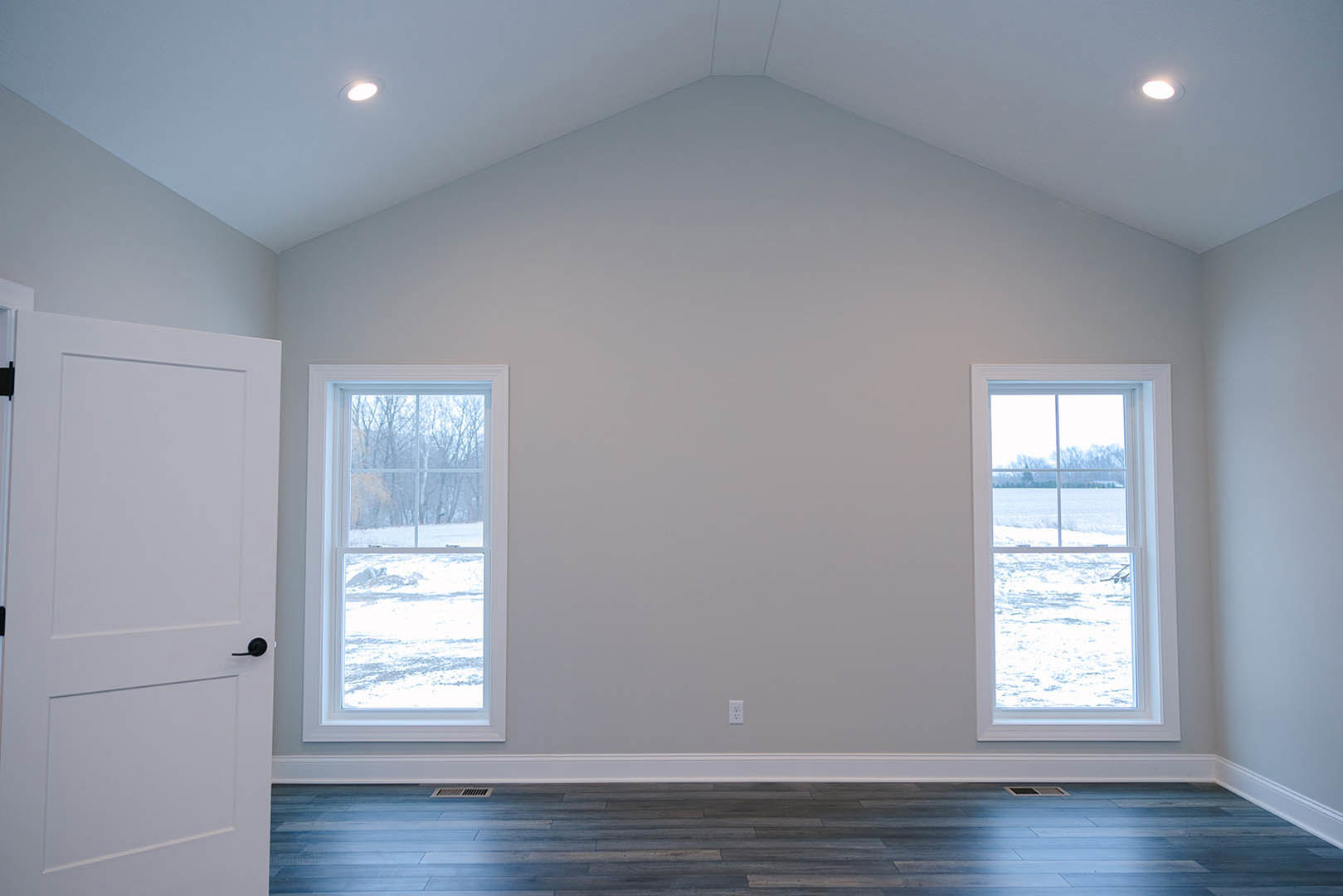 White-painted room with two large windows overlooking a snowy field, wood flooring with white baseboards, and a white door featuring a black handle.
