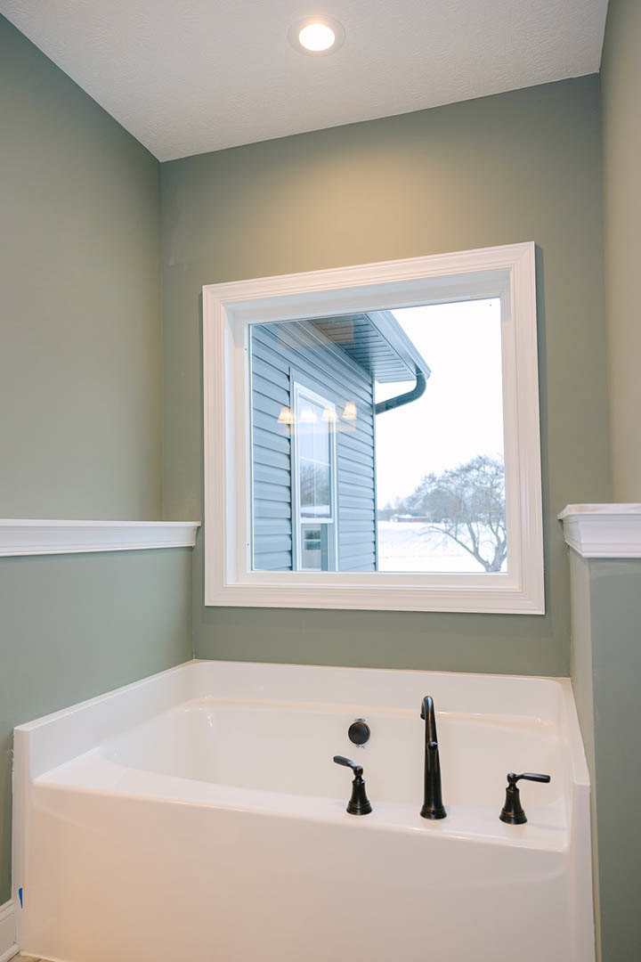 Bathroom with white-framed window, built-in bathtub with chrome faucets, white sink, green accent wall, ceiling light, and white shelving.
