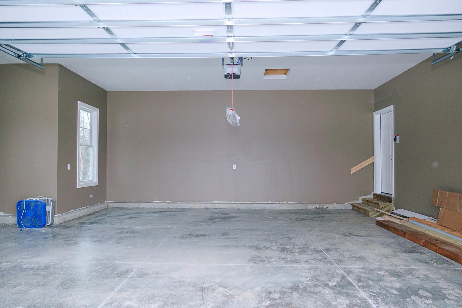 Concrete-floored garage with white-framed window, white door in wooden frame, exposed metal framing and wiring, blue and white ceiling fan, plaster walls, and staircase leading