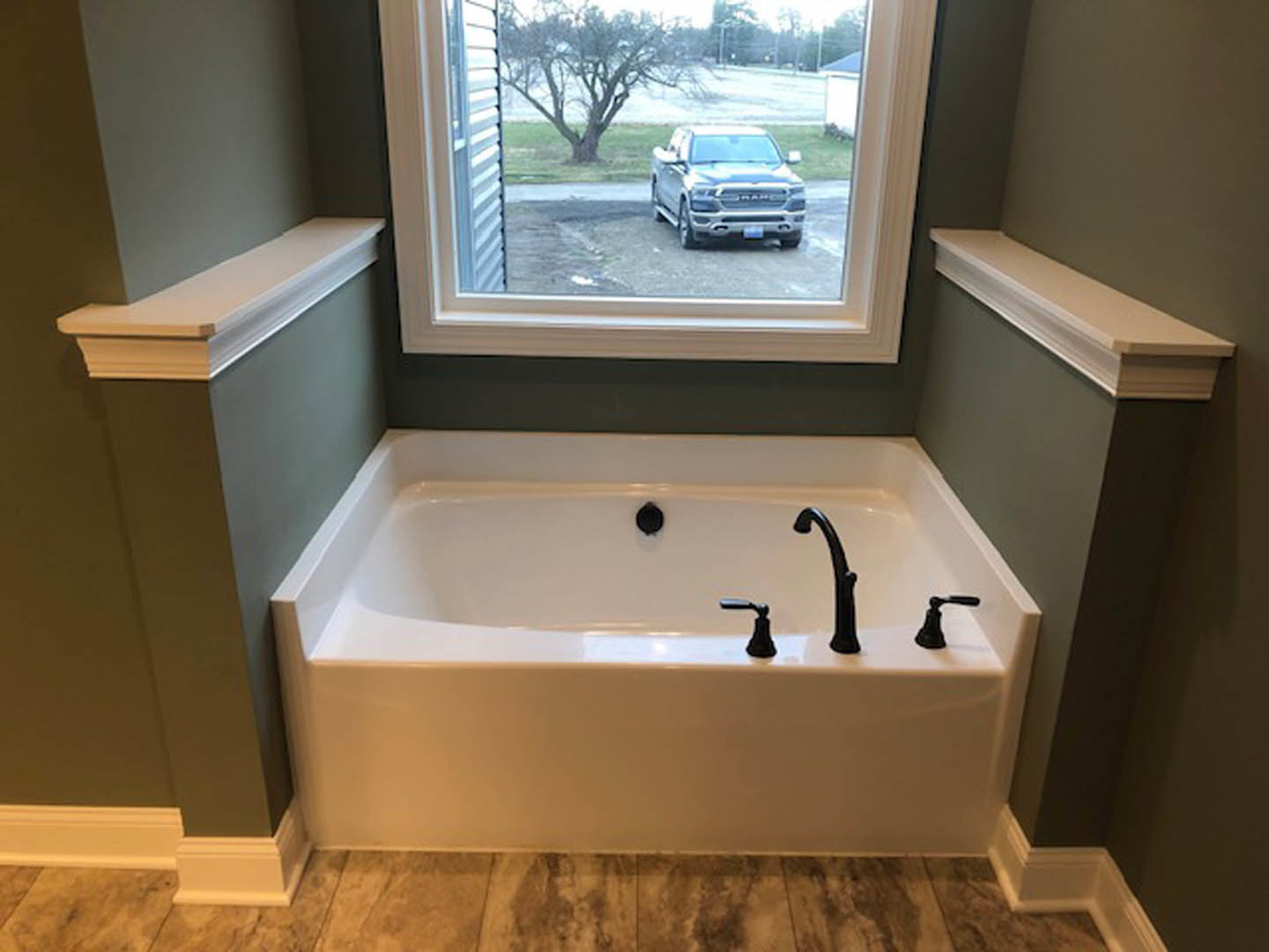 Freestanding bathtub with chrome faucet beneath large window, silver truck visible outside on driveway, white walls and bathroom accessories