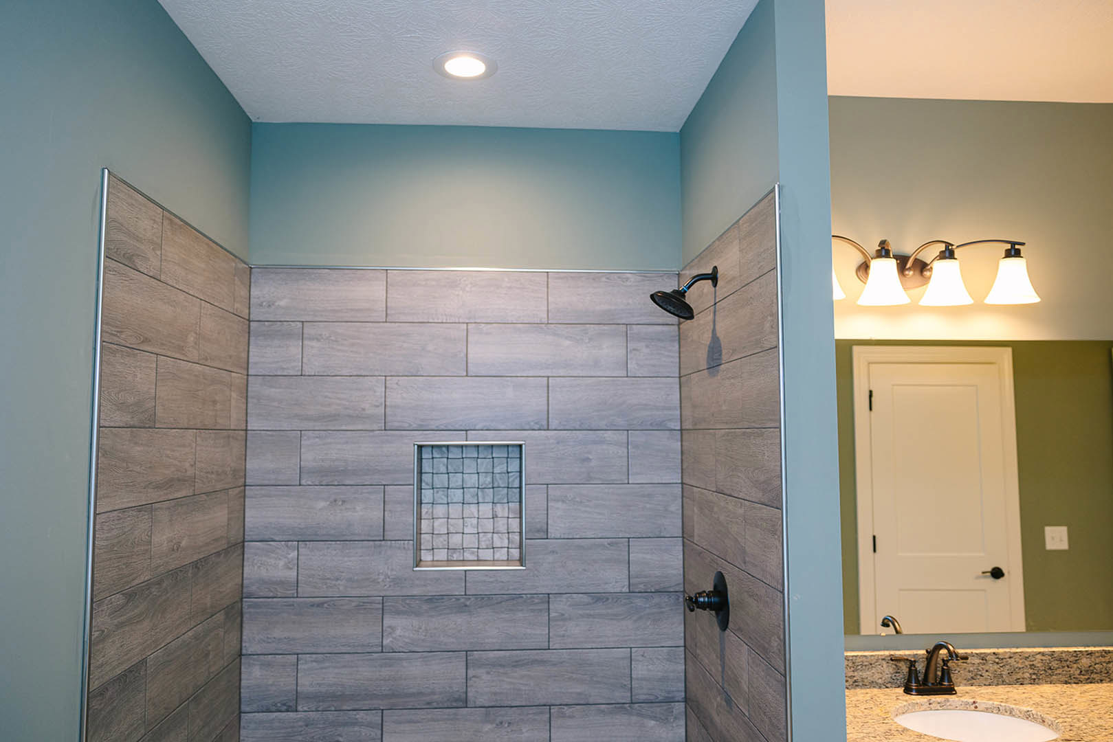 Walk-in shower with gray tile wall, white door featuring black handle, grid-patterned tile detail, modern sink, brushed metal light fixture, recessed ceiling light, tile flooring