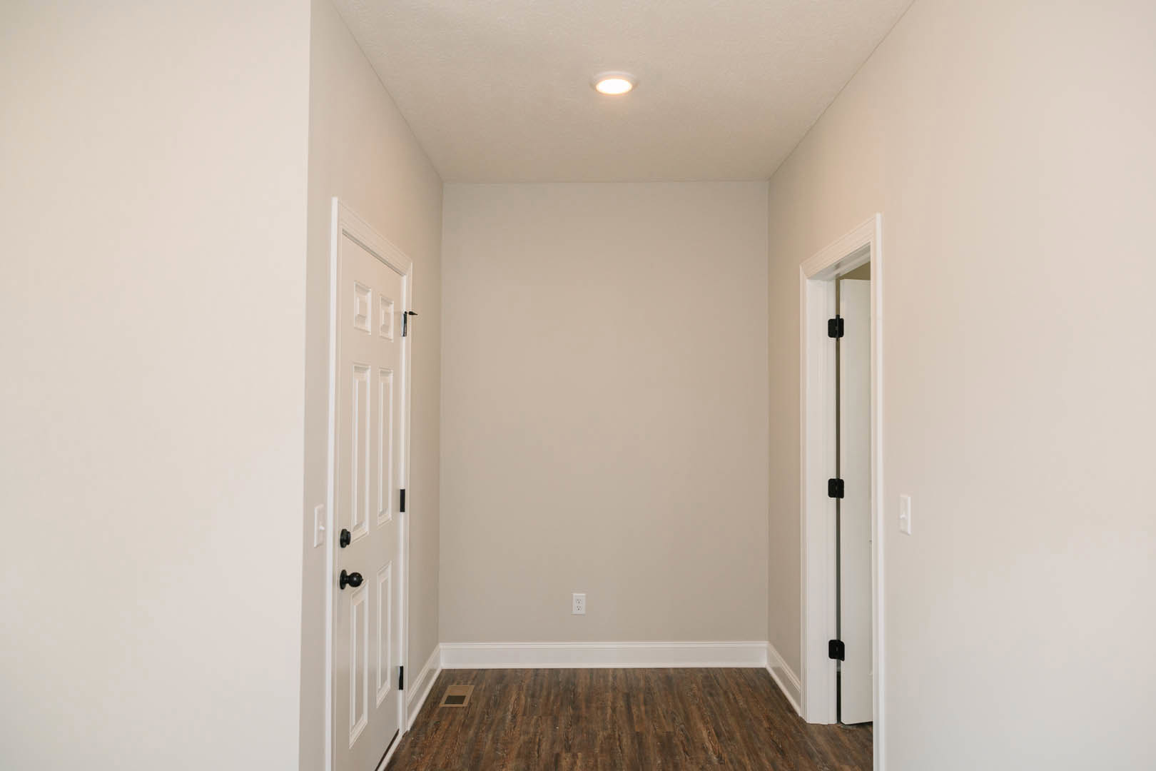 Hallway with wood laminate flooring, white baseboards, two white doors with black hinges, white plaster walls, ceiling light, and soft shadows on the wall