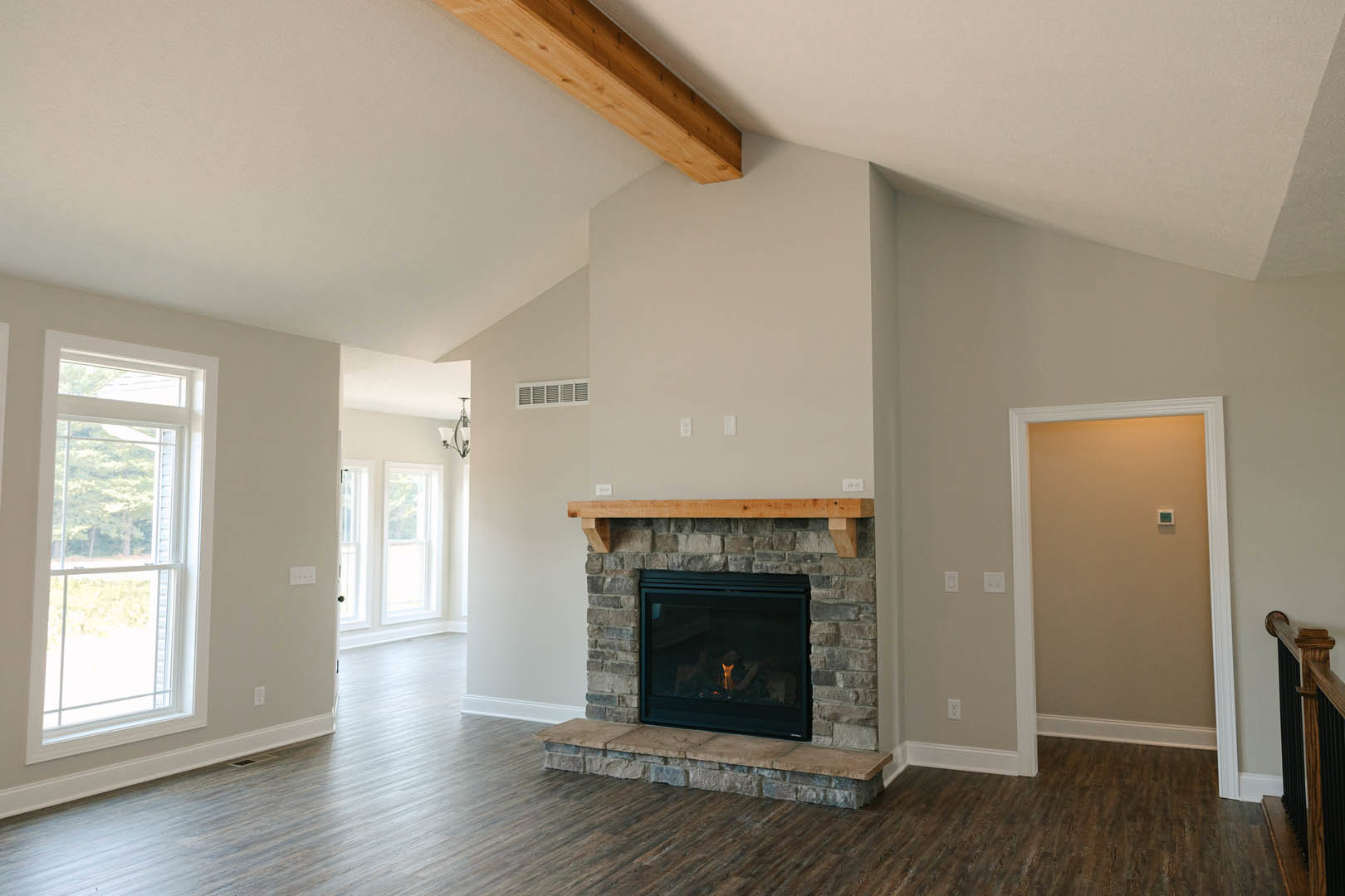 Stone fireplace with burning fire, wood flooring, large window overlooking trees, wood beam on brick wall, light switch on adjacent wall