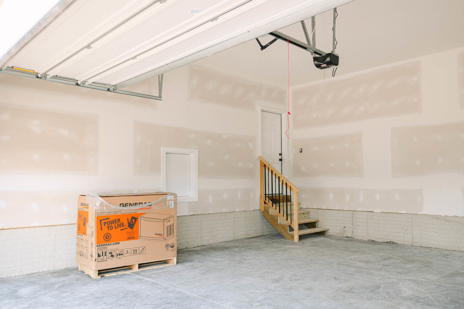 Garage interior with plaster walls, wooden staircase featuring black railing, large cardboard box on a pallet, white door with matching frame, and black object with red label on