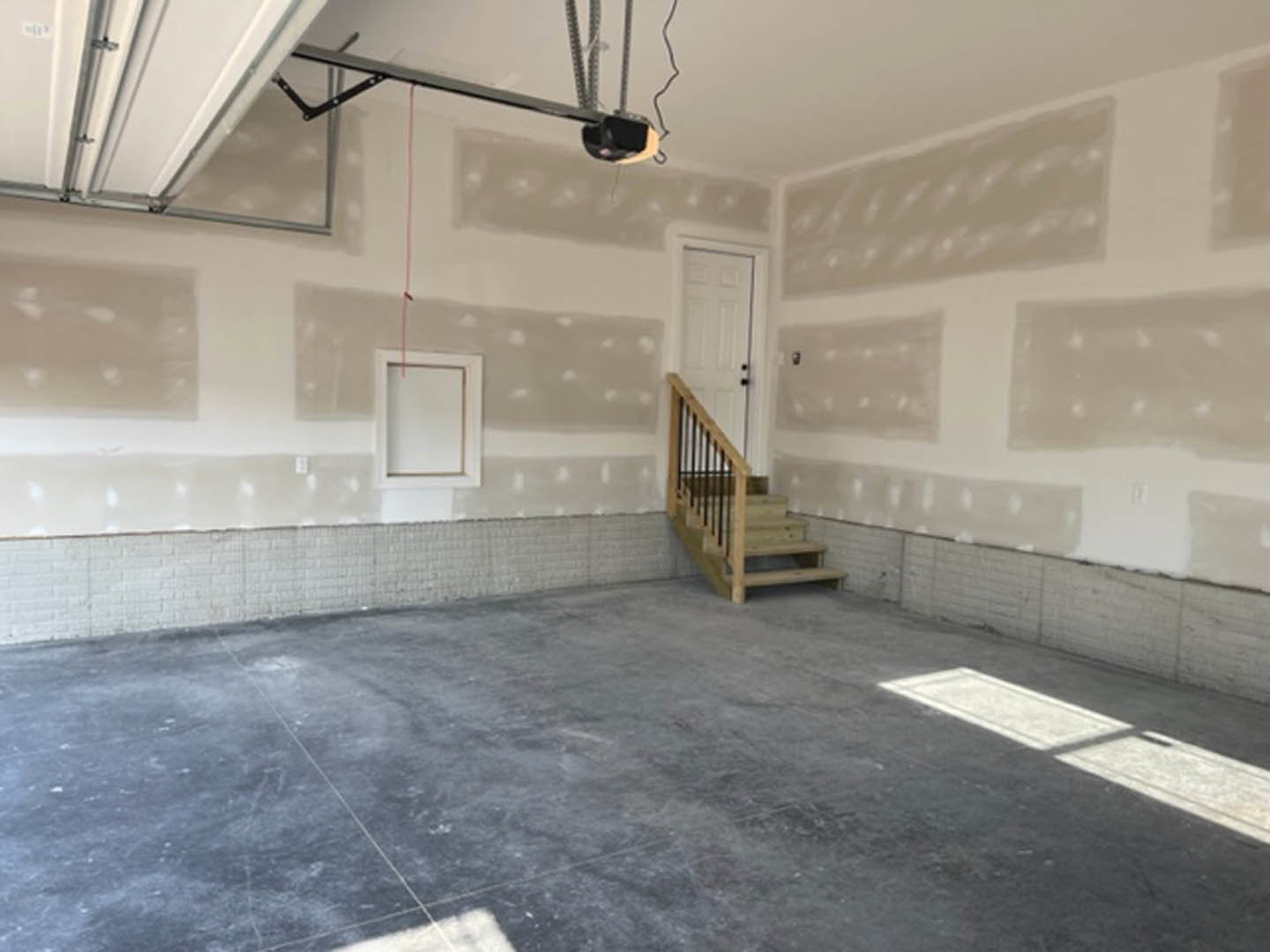 Wooden staircase with black metal railing, concrete floor, white door with black knobs, white walls, shadow cast across floor, red tape on door frame
