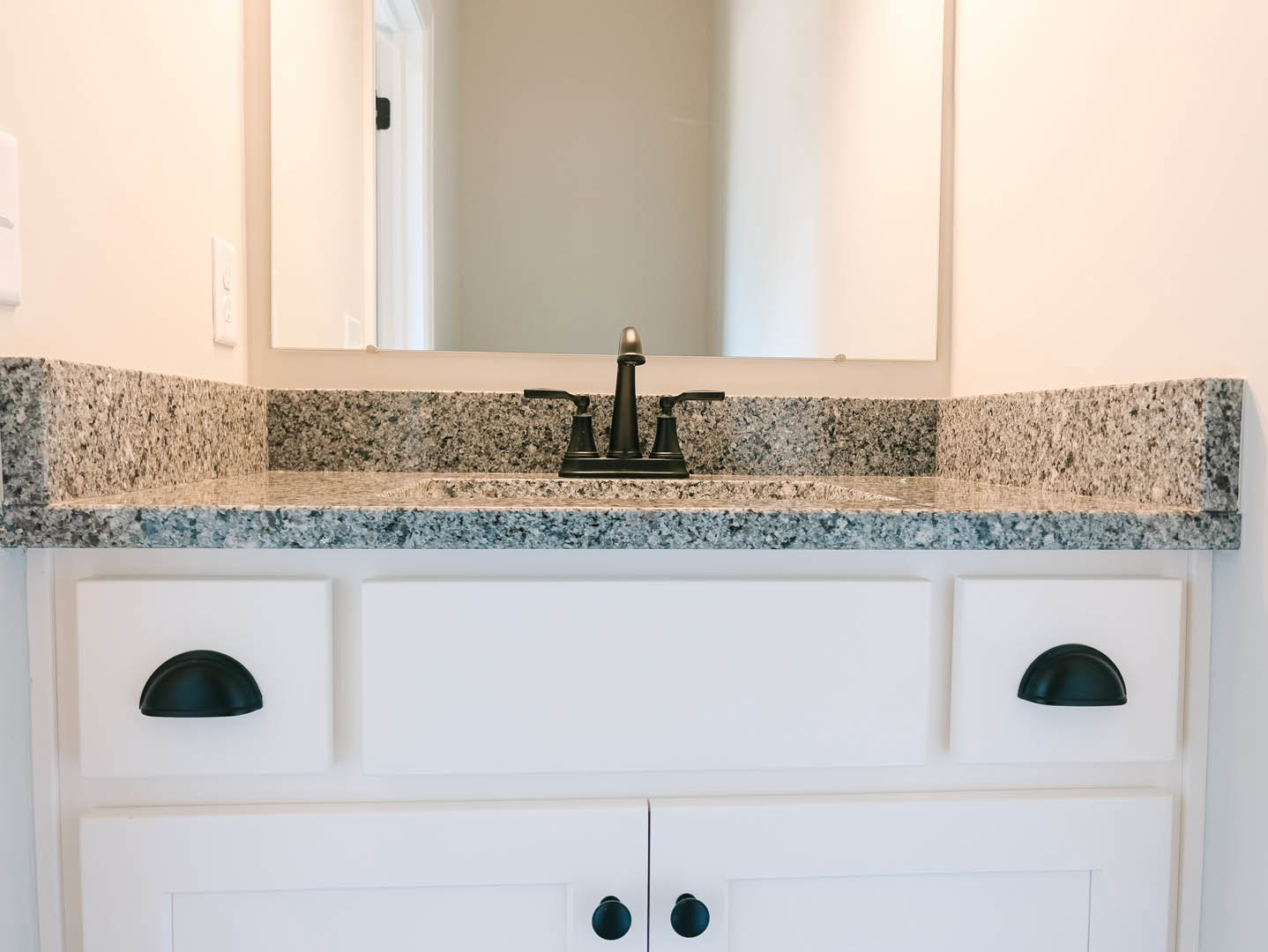 White bathroom vanity with black handles, rectangular sink, chrome faucet, and large framed mirror mounted above on light-colored wall