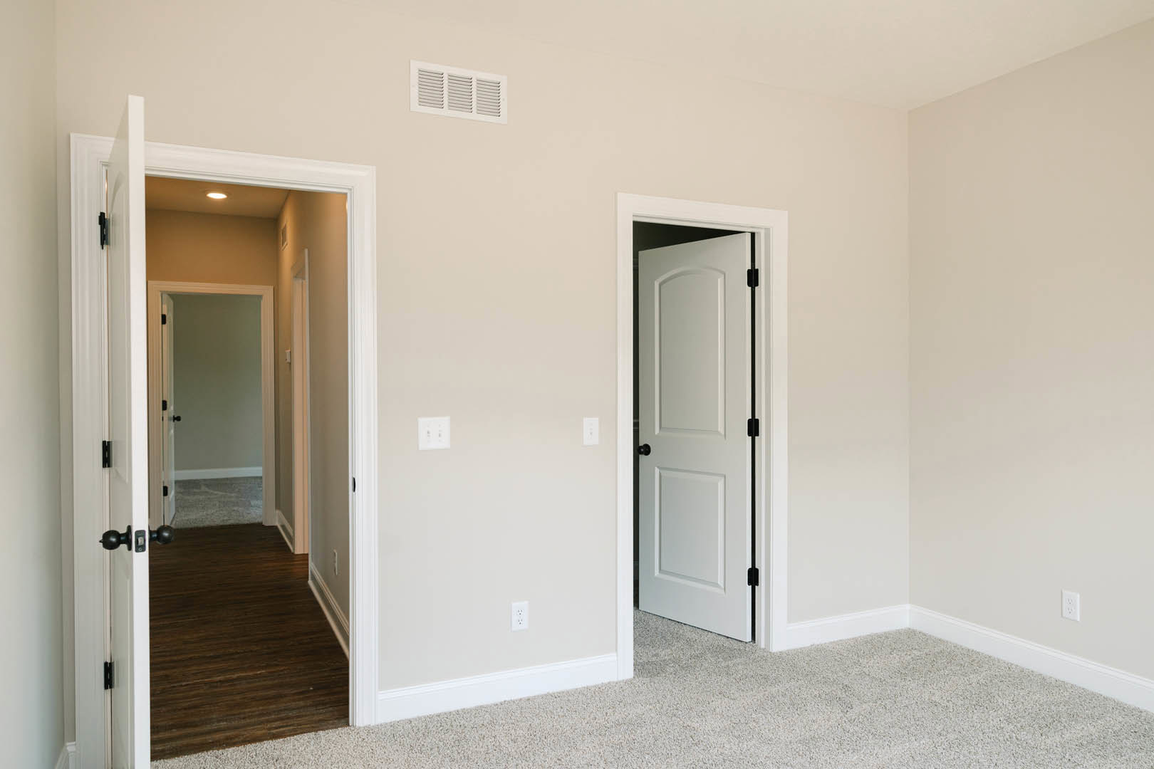 Open white door leading to hallway with dark wood flooring, white walls, ceiling vent, wall outlet, and black light fixtures
