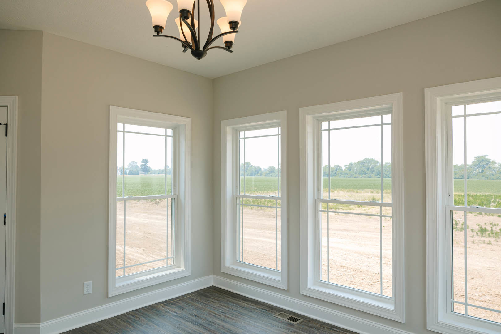 Bright room with three large windows overlooking a grassy field, wood flooring with white baseboards, modern chandelier hanging from ceiling, blue accent object mounted on white