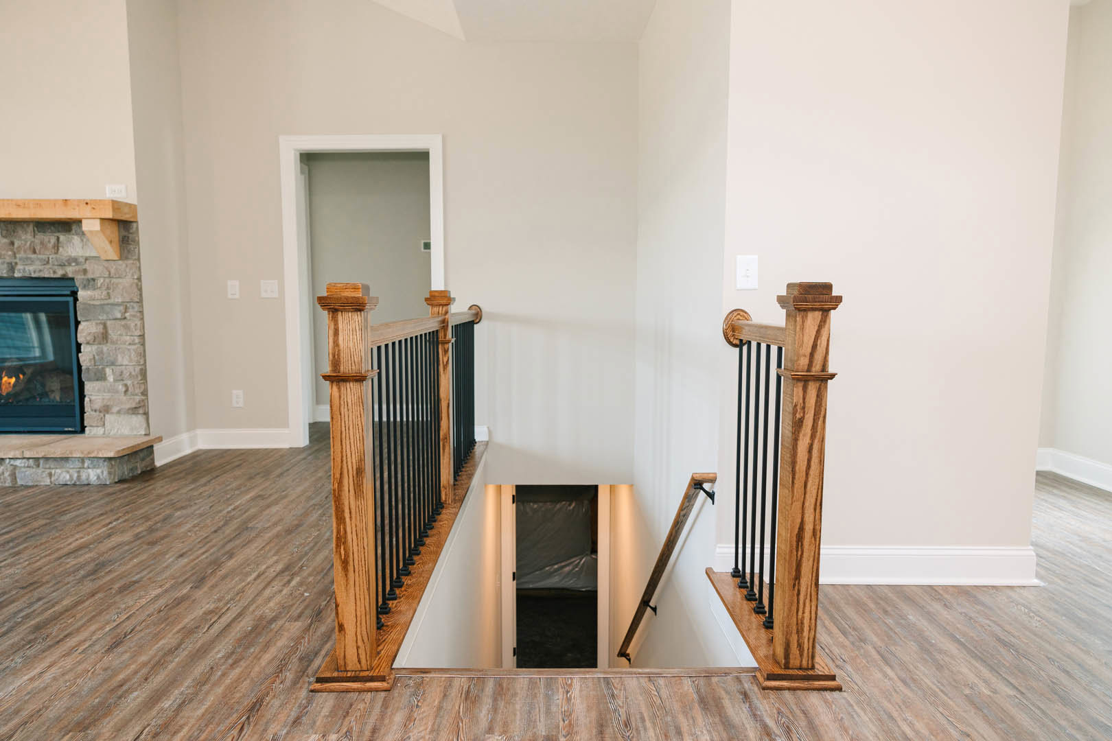 Hardwood staircase with metal railing leading to a white door, stone wall fireplace with visible flames, wood flooring throughout, plastic bag hanging on door handle