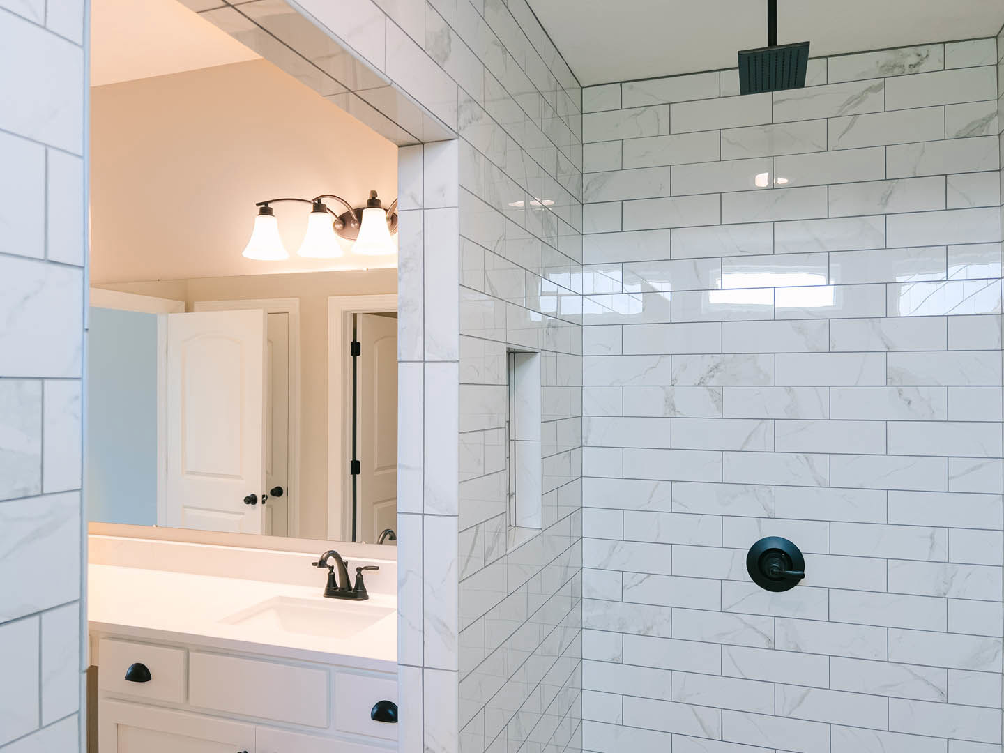 White bathroom with rectangular sink, wall-mounted mirror, three-light fixture above, white cabinet door with chrome handle, white tile backsplash featuring black accent line