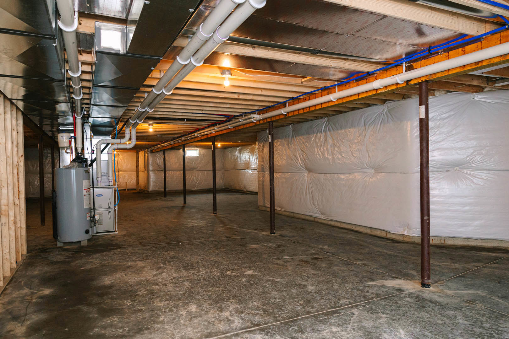 Basement utility room with exposed ceiling pipes, large white water heater, white plastic wall covering, concrete floor, and metal support poles
