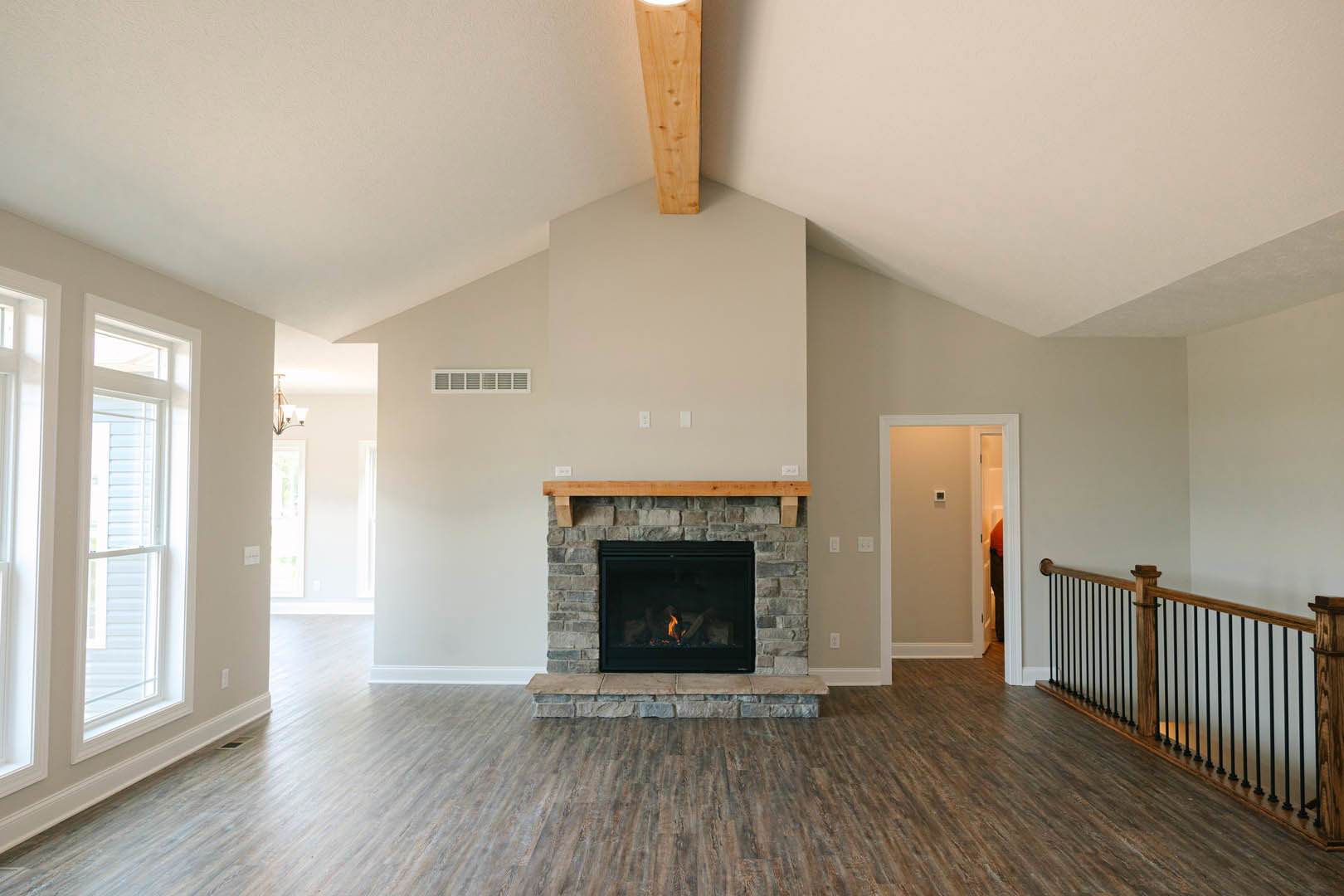 Stone fireplace with burning fire, wood flooring, black metal railing, white plaster walls, window with trim, and door with light switch in modern living room