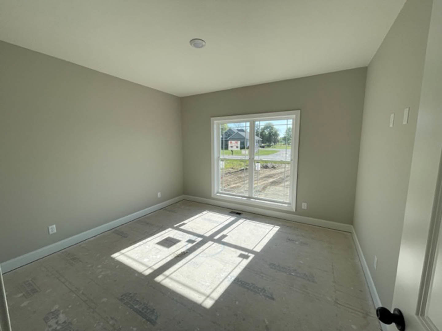 Sunlit room with large window overlooking road and grassy field, tile floor with window shadow, white circular object on white surface near wall.