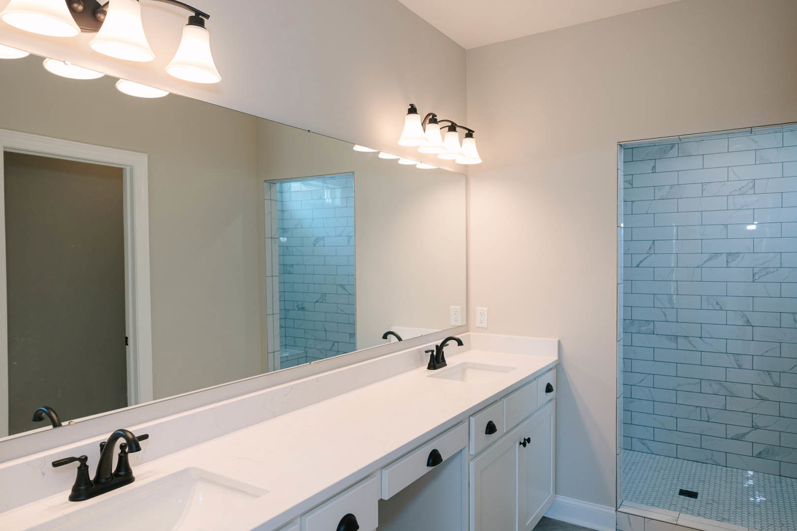 Bathroom featuring a wide mirror above white countertops, white tile shower with wall-mounted light fixture, and sleek cabinetry.