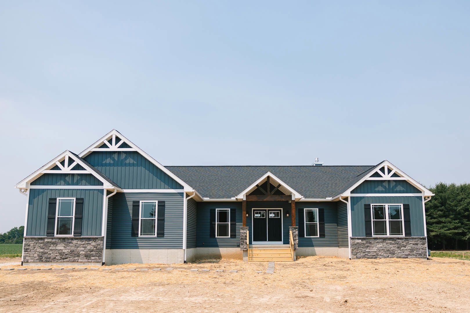 Blue metal roof atop a cottage-style home with double glass doors, white-framed windows, covered porch, and landscaped front yard with a tree and dirt field.