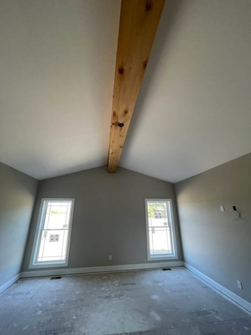 Living room with exposed wood ceiling beam, plaster walls, two large windows, light hardwood floor, and a close-up view of a paneled door.