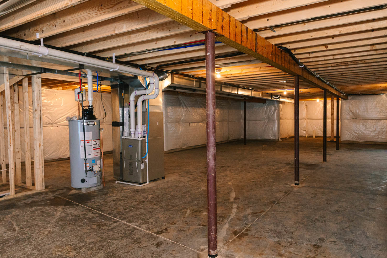 Exposed steel pipes and beams in an unfinished basement with a water heater, wooden support pillar, and partial view of a couch on concrete flooring.
