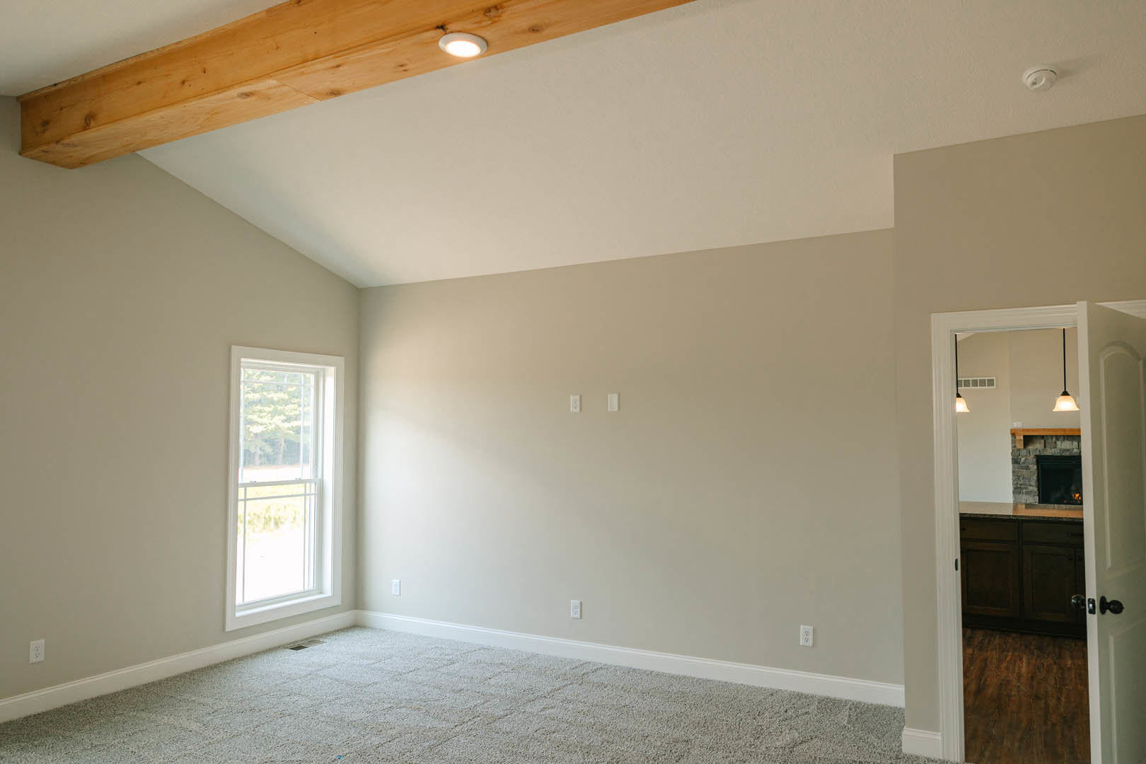 Carpeted room with white-framed window, wooden ceiling beam, recessed ceiling light, and door opening to a kitchen