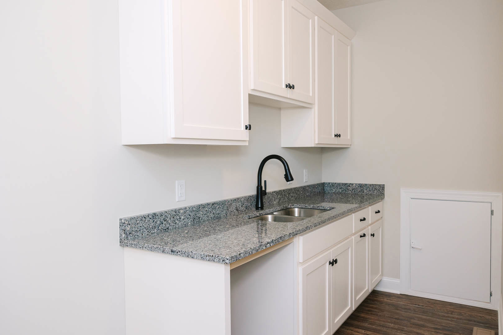 White kitchen cabinets, granite countertops, black faucet with white sprayer, wood flooring with white trim, white door featuring metal handle and hinge, stainless steel sink