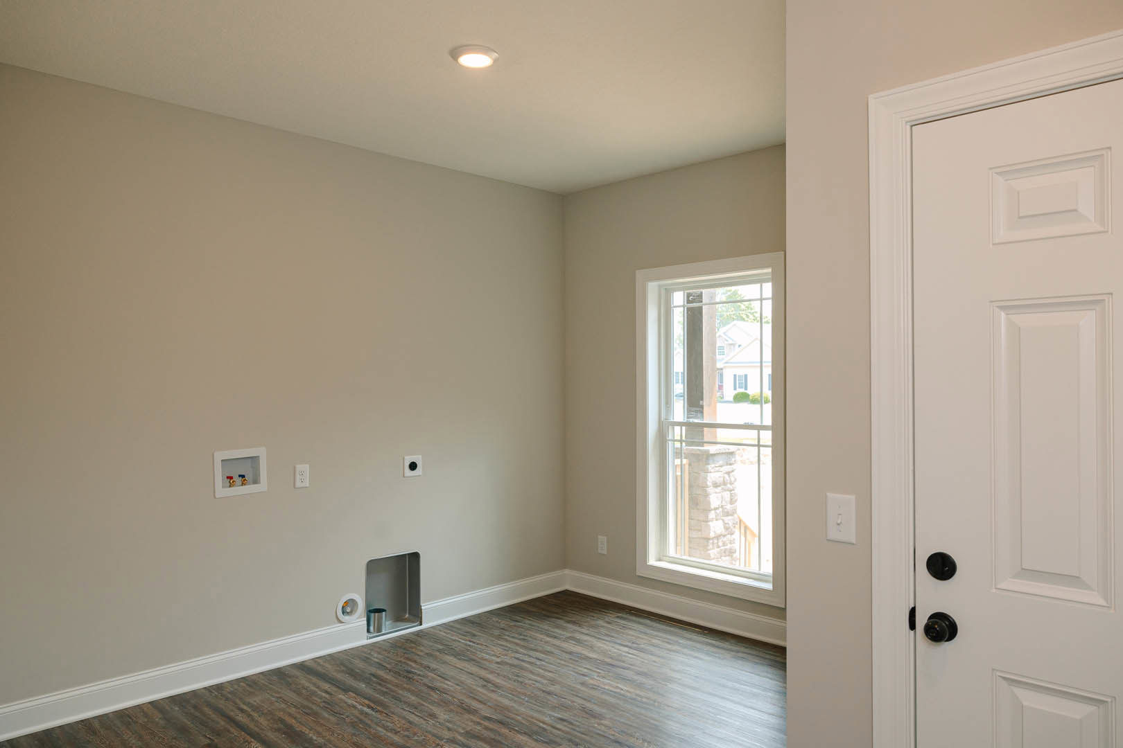 White paneled door with black knob, wood laminate flooring, white walls with base molding, window showing neighboring house, metal trash can near door, light switch on wall