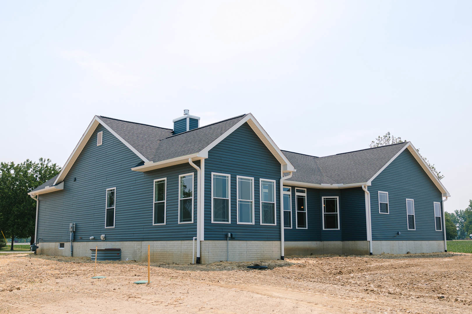 Partially built house with blue siding, white-framed windows, exposed gutters, dirt construction site, and clear blue sky