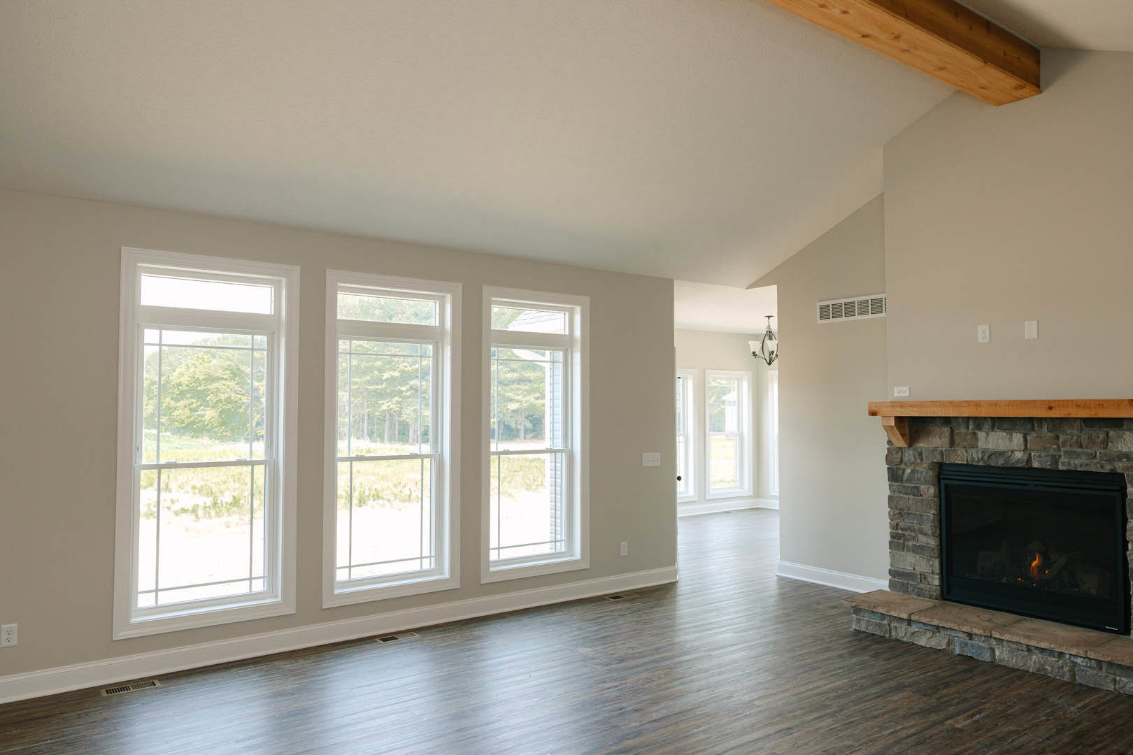 Living room with hardwood floors, white-trimmed windows overlooking a field and trees, stone fireplace with active fire, neutral walls, crown molding