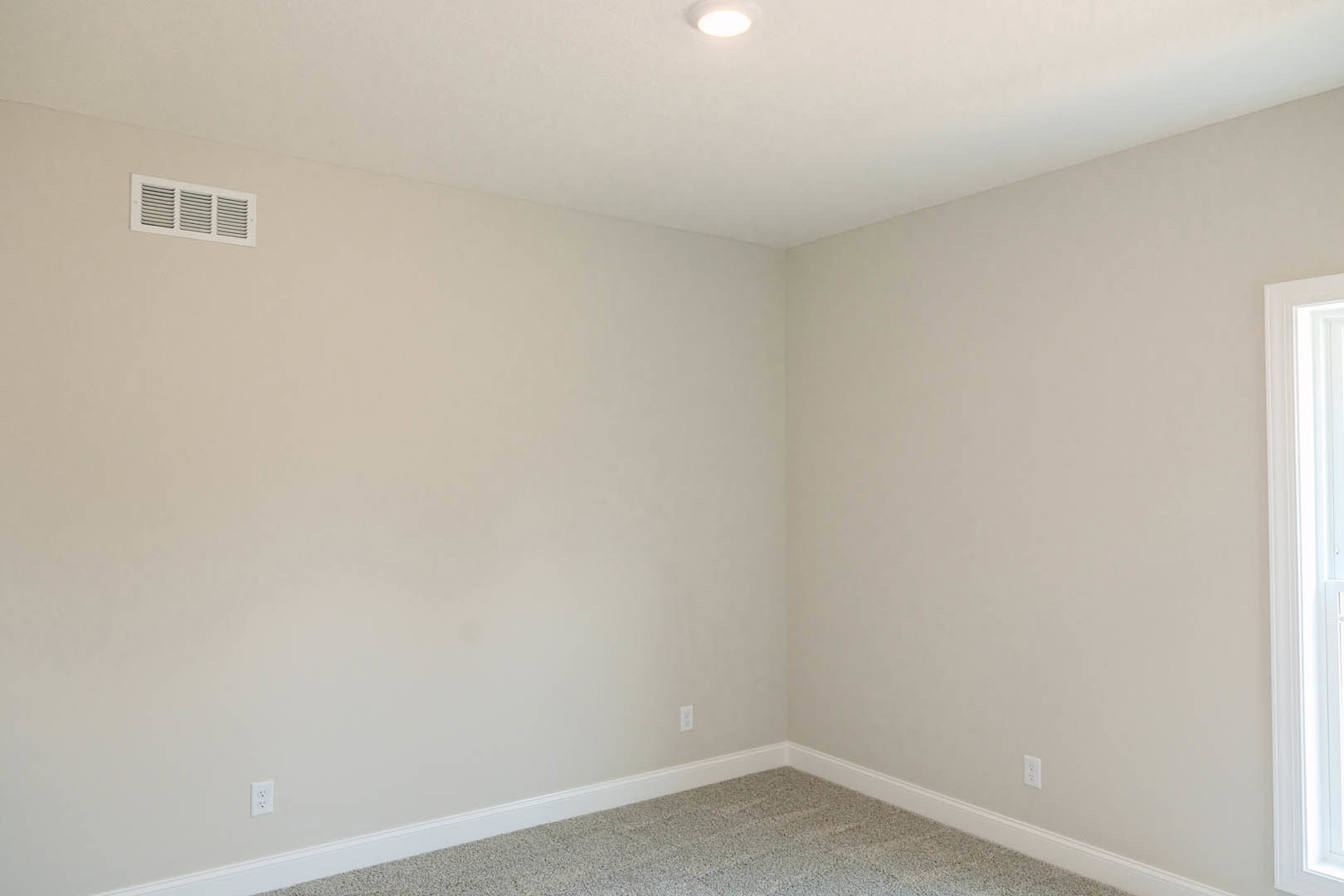 Corner room with white plaster walls, carpet flooring, ceiling light fixture, and vent installed in the upper wall near the ceiling molding
