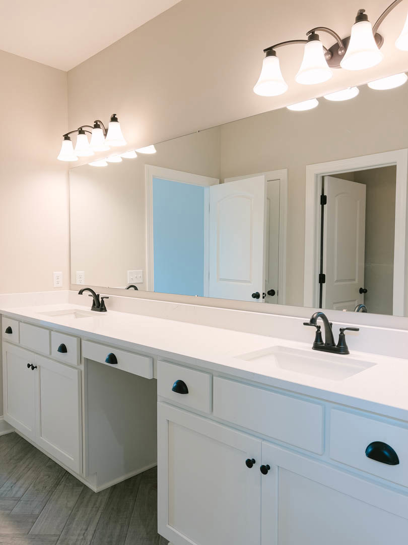 Bathroom with expansive mirror above white countertops, white cabinetry, matte black faucets, blue accent wall, and white door with black hinges.