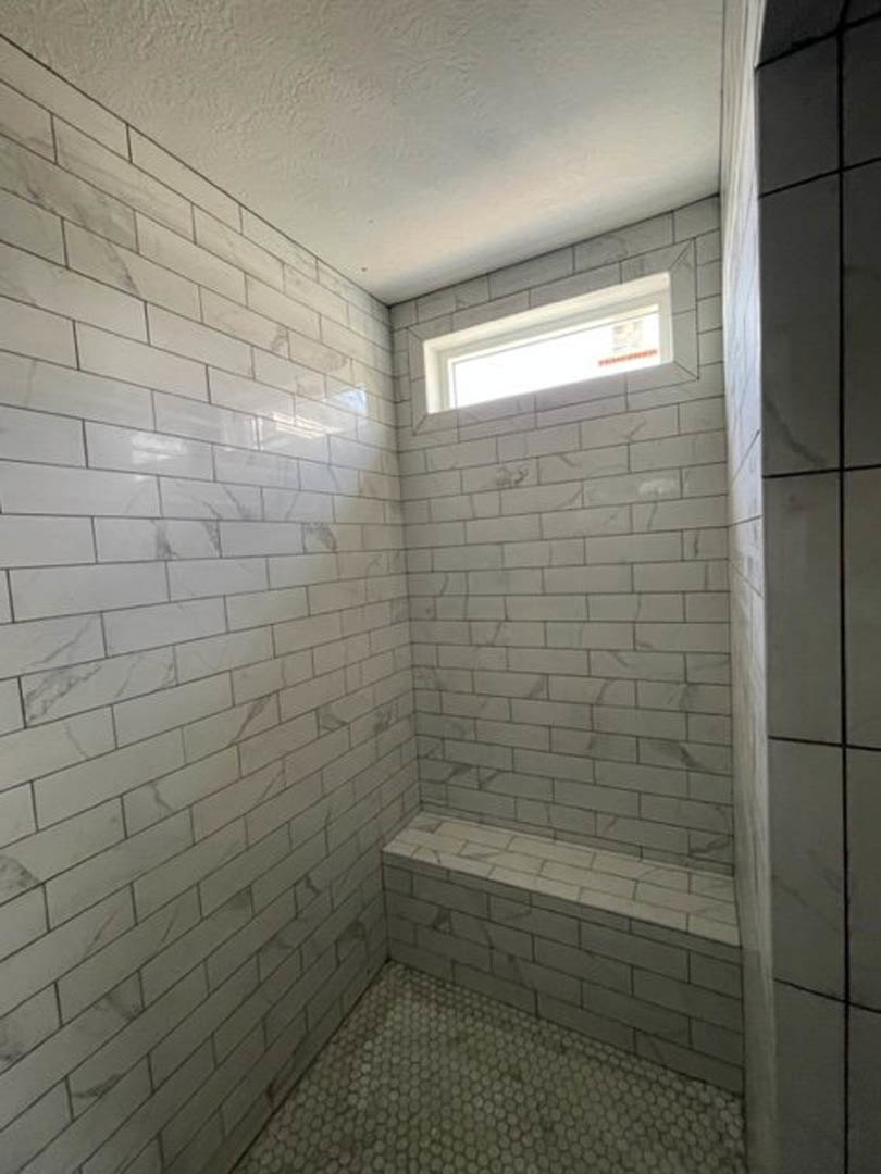Walk-in shower with large window, built-in bench, light fixture mounted on exposed brick wall, stone tile flooring, and tiled walls