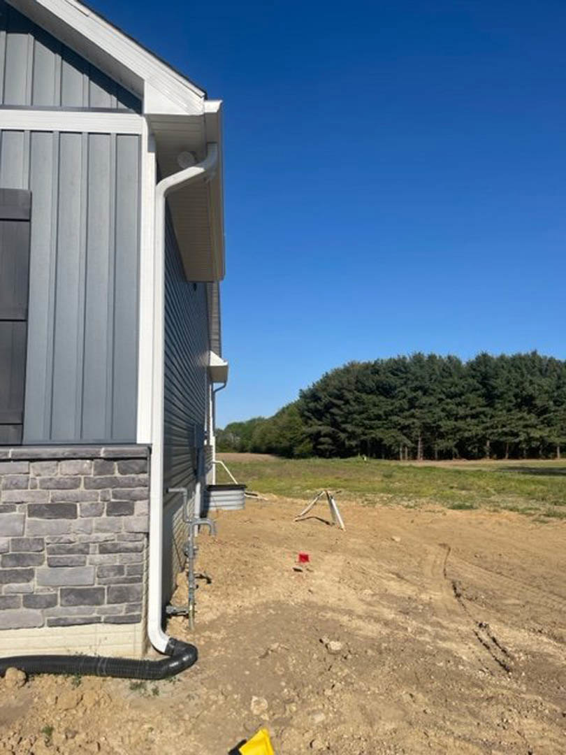 Two-story house with brick and siding exterior, large leafy tree nearby, dirt field in foreground, grassy area and additional trees in background