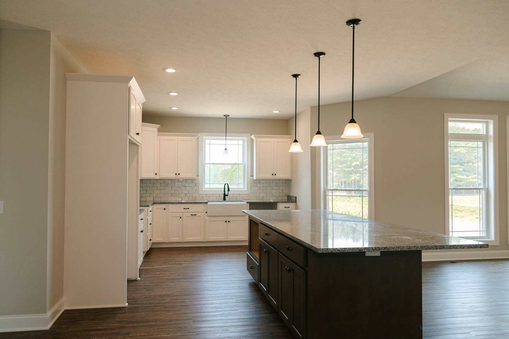Spacious kitchen featuring a large granite island, hardwood flooring, white-framed window above a sink, modern light fixture, and black support pole