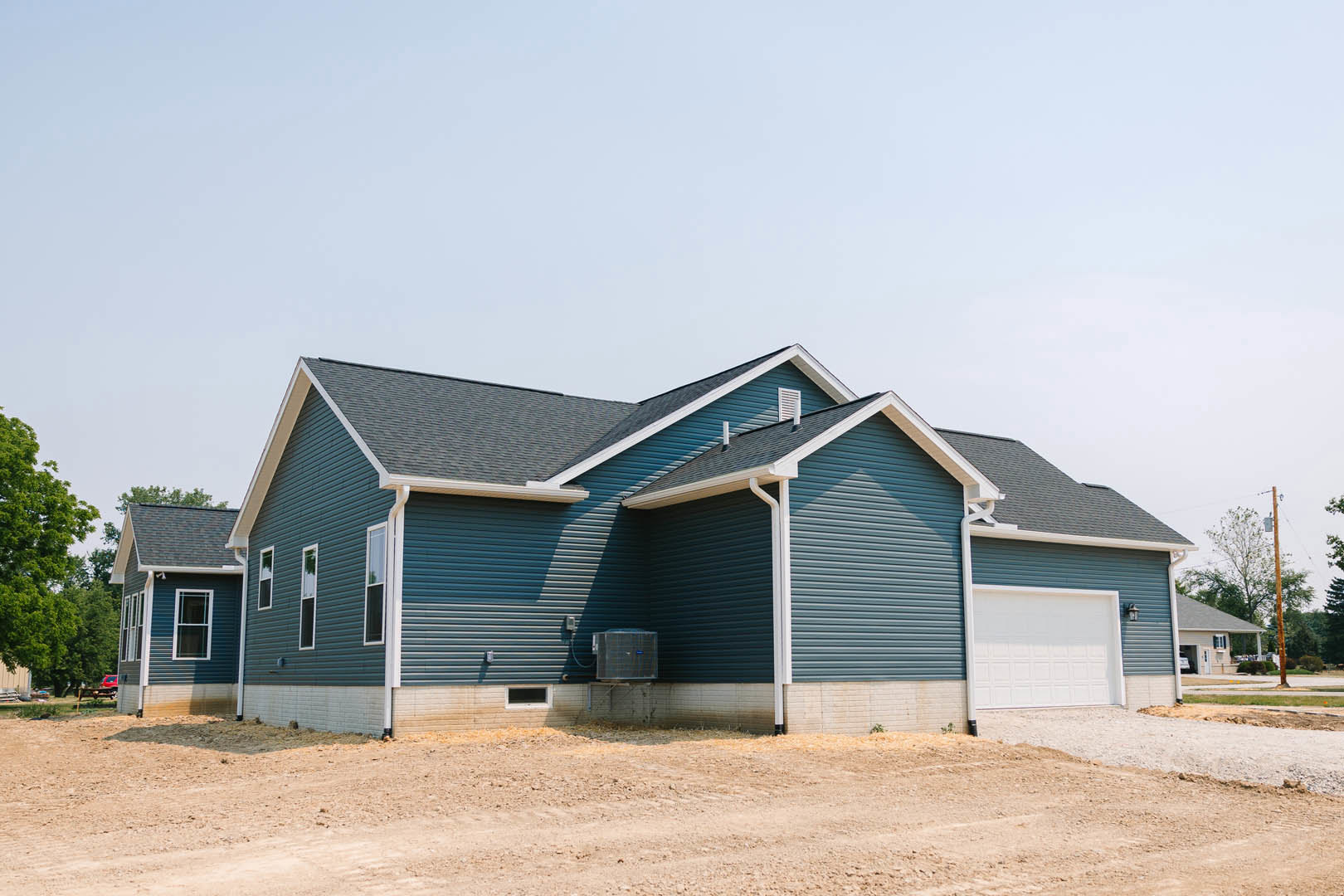Blue siding house with white roof, attached garage, large front window, mature tree in foreground, person standing near garage door, dirt yard bordered by brick wall