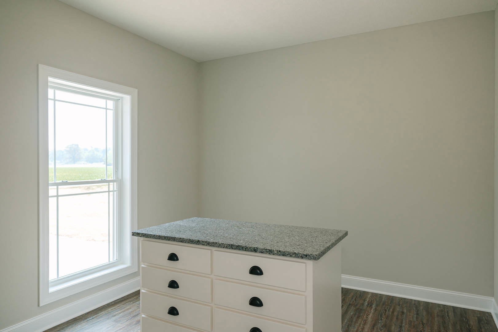 White cabinetry with black handles beneath a white countertop, wood flooring, large window overlooking a grassy field
