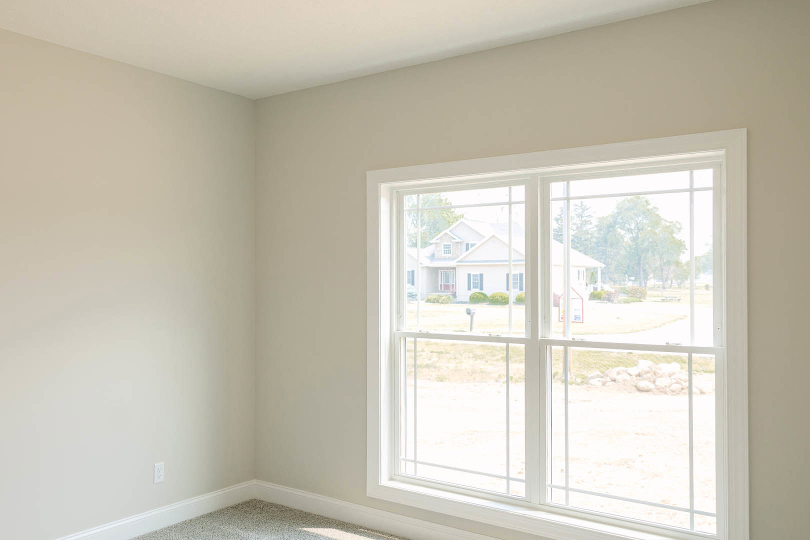 Spacious room featuring a large window with white trim, offering views of a grassy field and neighboring house; light plaster walls and floor visible, window blinds partially