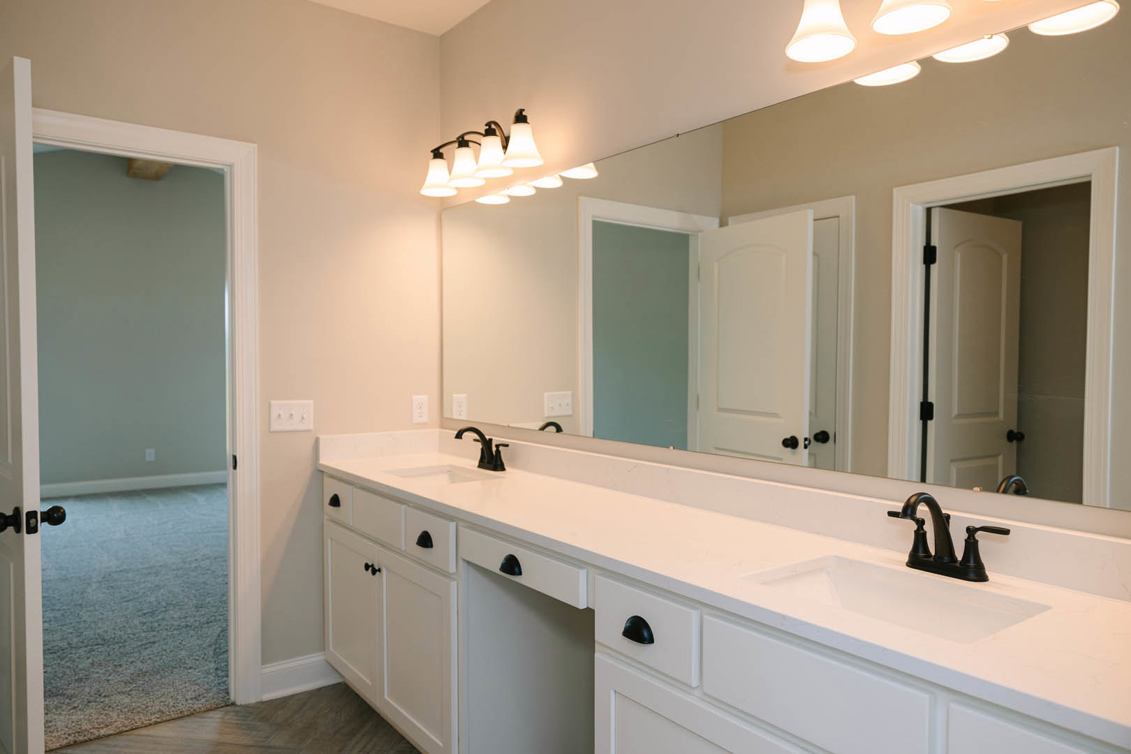 Bathroom with expansive mirror above double sinks, white countertop with black handles, white cabinetry, light switch on wall, white door with black knobs, ocean view visible