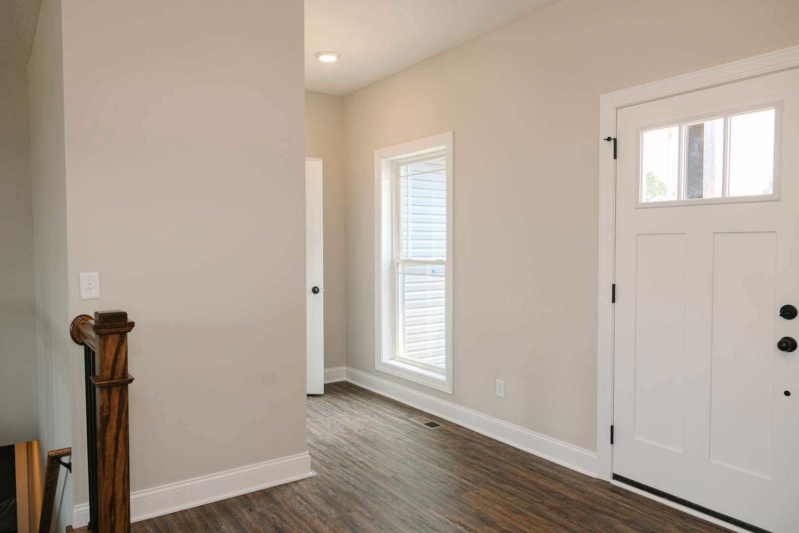 Hallway with light laminate wood flooring, white walls, white door with black knobs, window set in white trim, wooden box with black top, and wall-mounted light switch