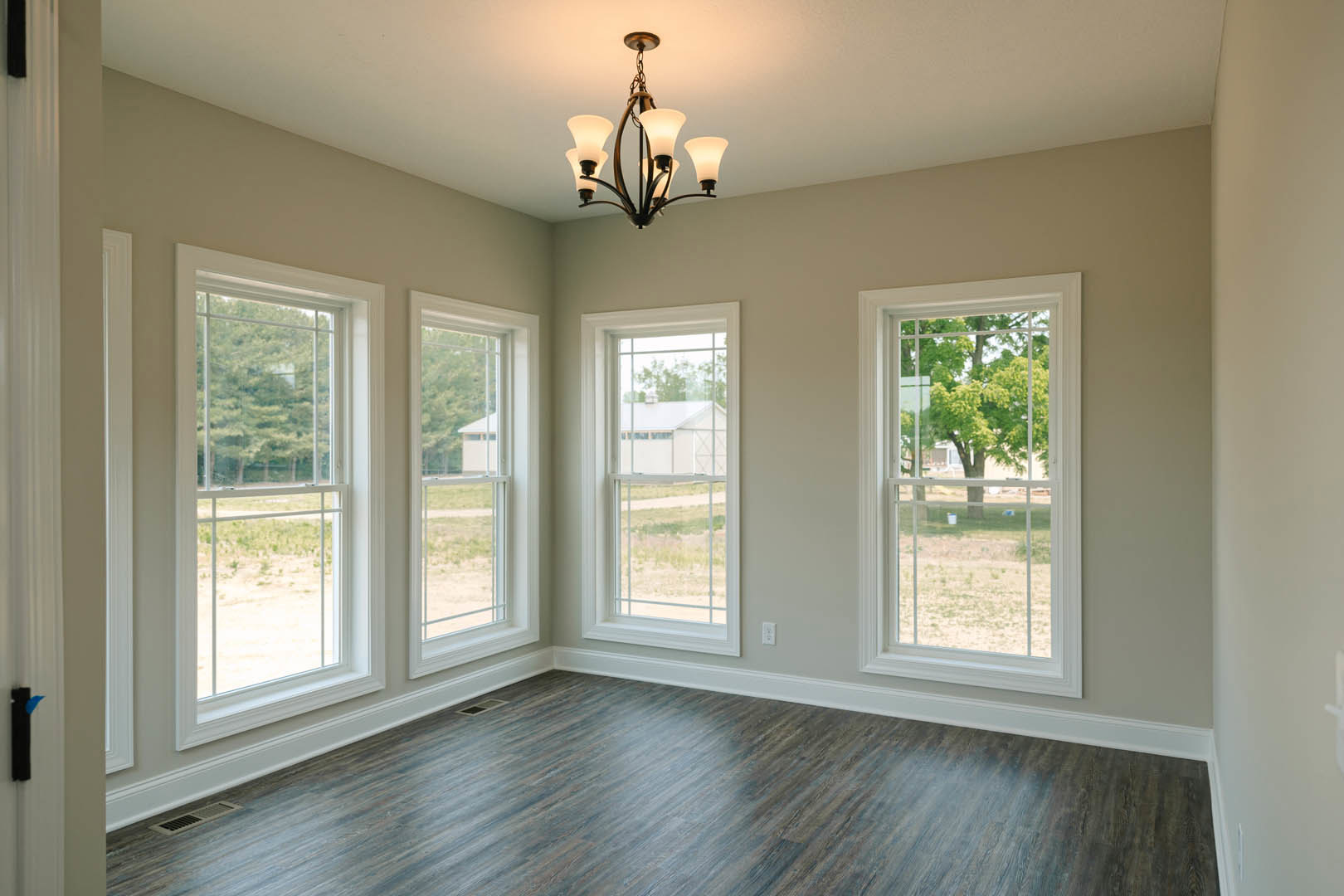 Chandelier hanging in a bright room with multiple windows, wooden flooring, white baseboards, and views of trees and a neighboring white house outside