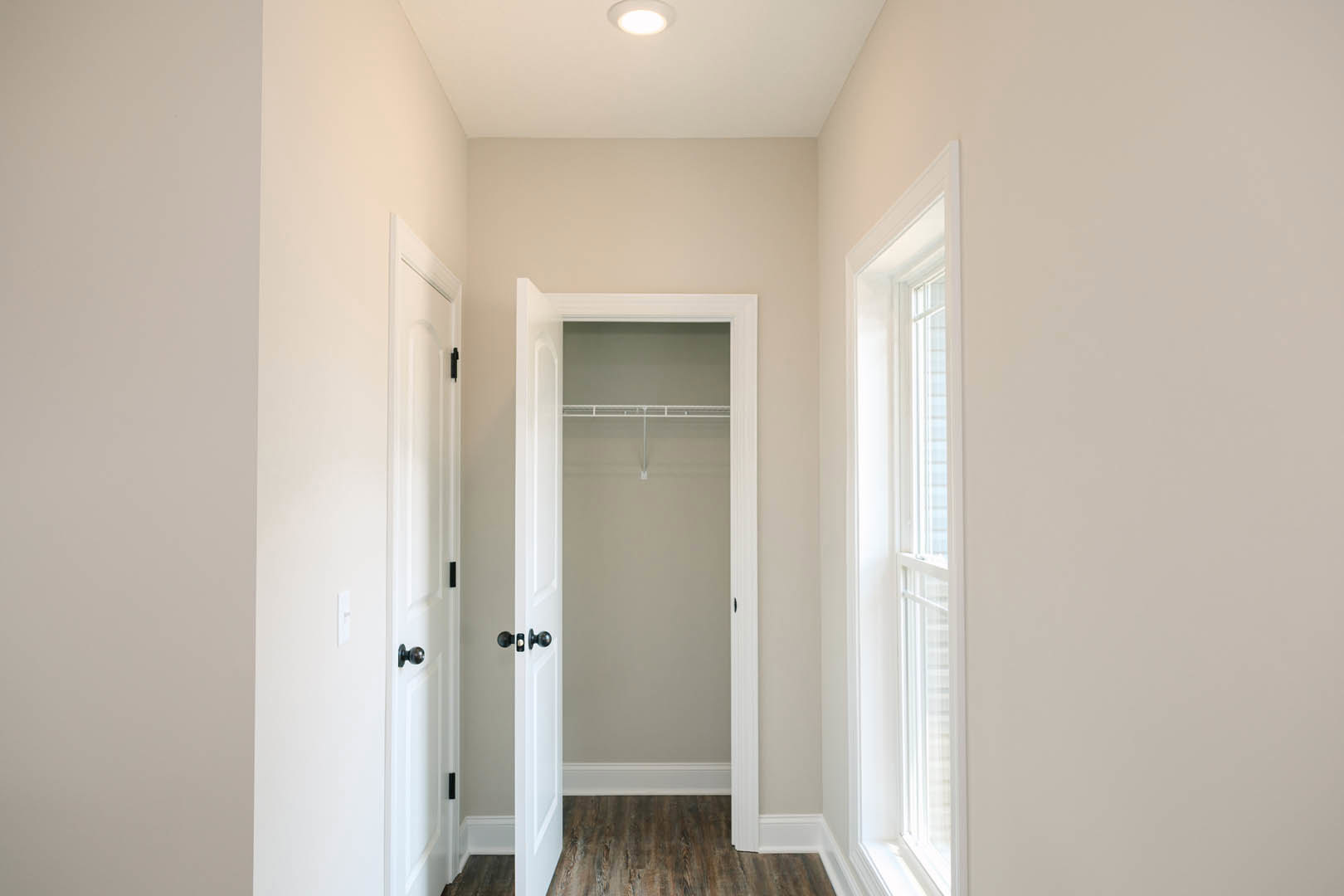 Hallway with white paneled doors, black handles, wood flooring, white walls, and ceiling, featuring a closet and window at the end