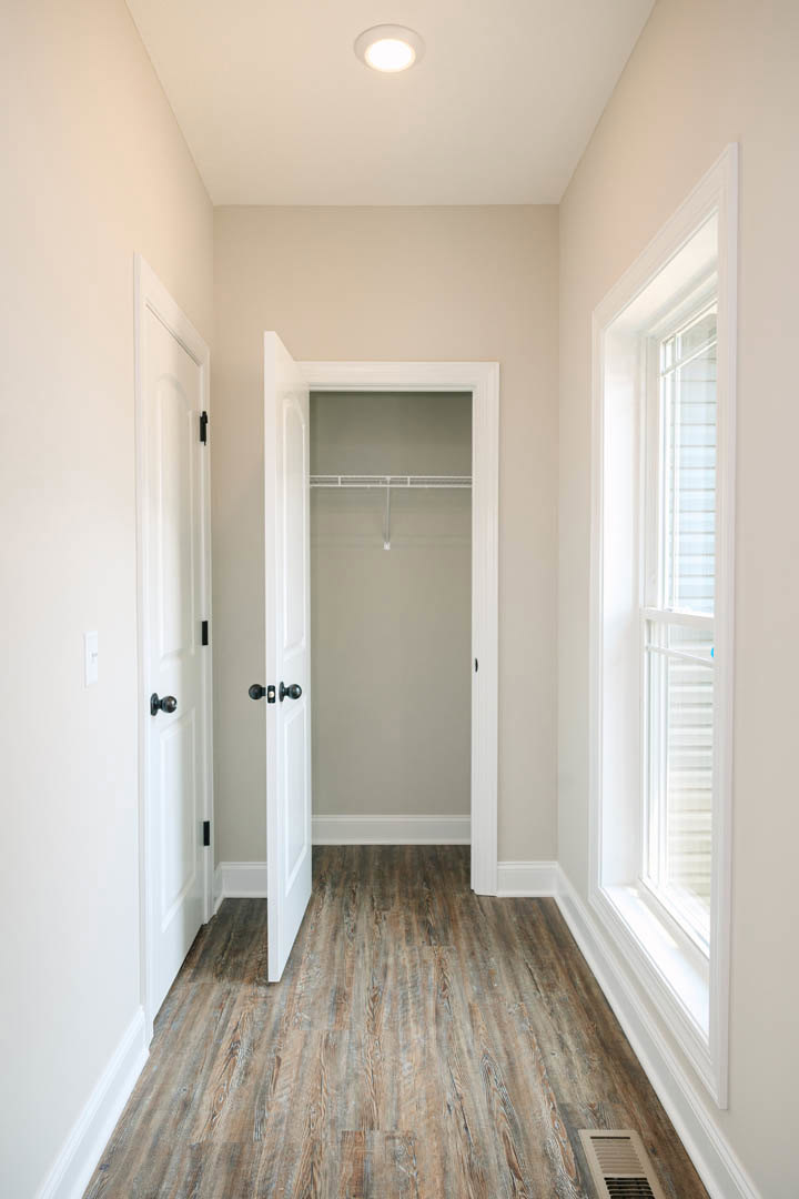 Open white closet door revealing wood flooring, wall vent, ceiling light, and close-up window with molding.