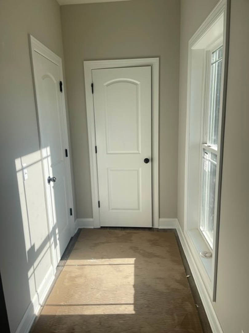 Hallway with brown carpet, white walls, two white doors featuring black and silver handles, window with white frame, simple molding