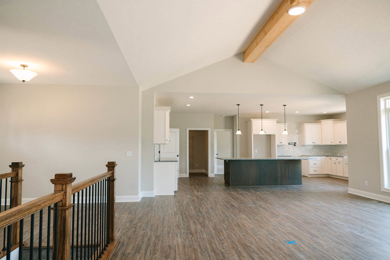 Spacious open floor plan featuring a central kitchen island, wide plank wood flooring, wooden railing with black metal bars, modern light fixture, and white plaster walls with