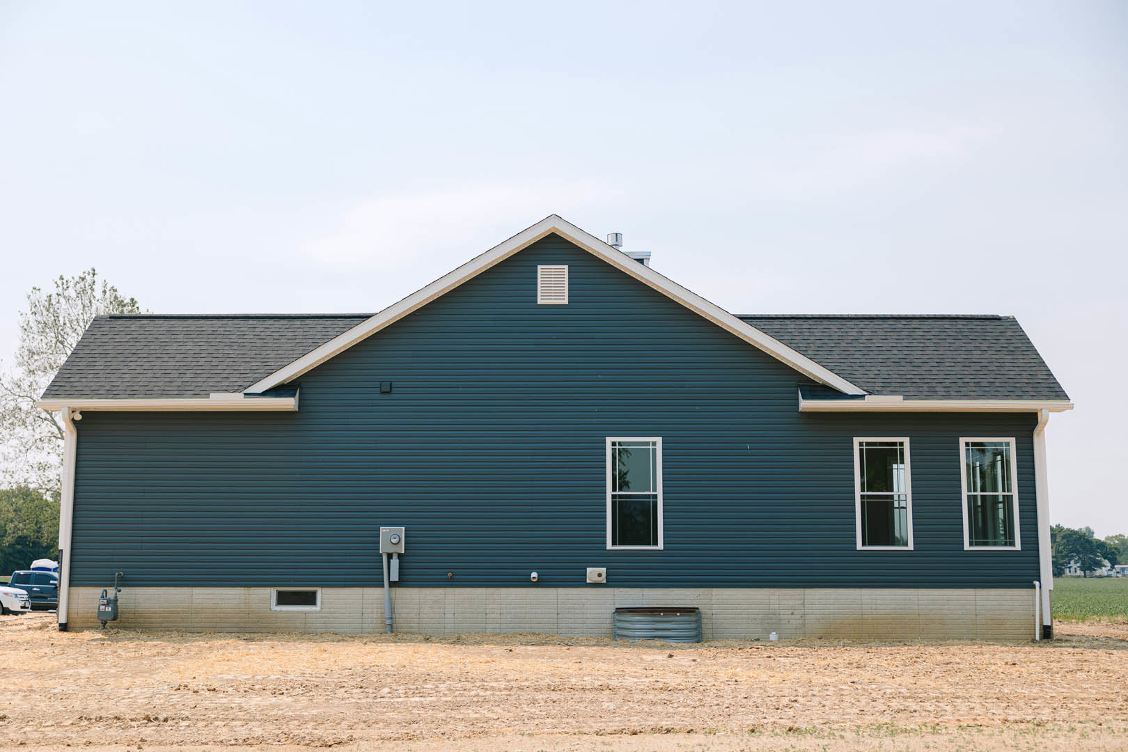 Blue horizontal siding on house exterior with white-framed window and white vent, dirt field in foreground, clear sky above